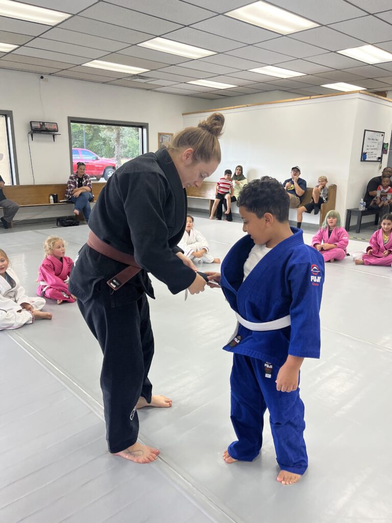 male child in blue kimono receiving a promotion on his belt by brown belt instructor in a black gi parents and other children in background
