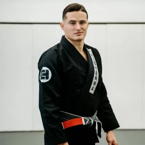 man in black gi and black belt posing for a photo in front of white wall mats