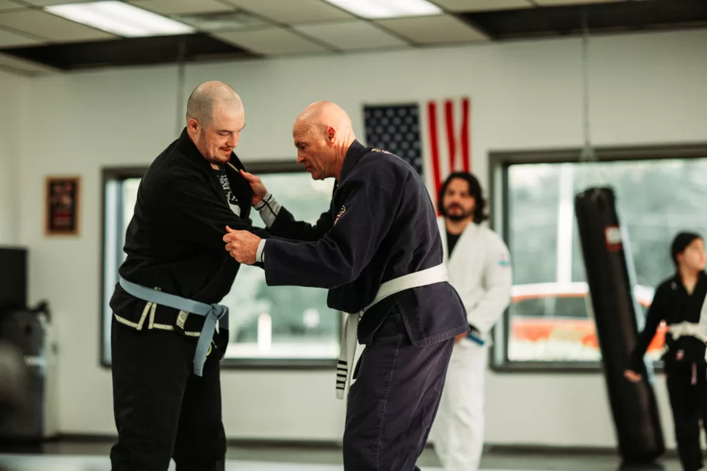 2 men in black gi's practicing judo techniques, man in white gi watching standing by windows and american flag hanging on the wall