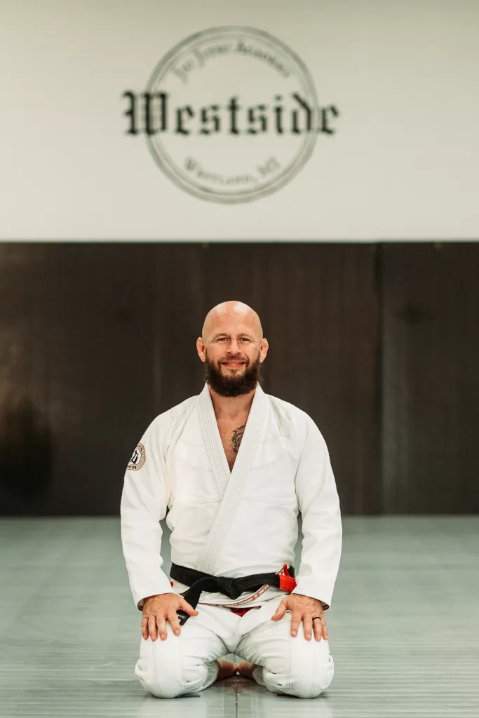 man in white gi and black belt kneeling on mat with westside jiu jitsu logo on wall above