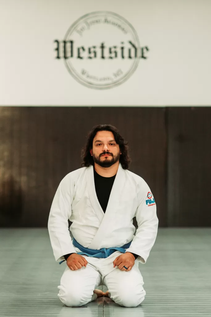 man in white gi and blue belt kneeling on mat with westside jiu jitsu academy logo abobe