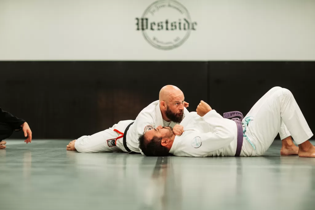 two men in white gi's lying on the grey mat black belt demonstrating a technique on a purple belt