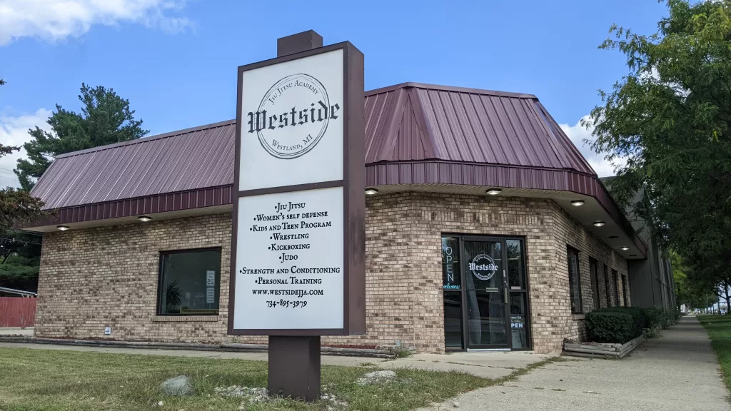 brown brick building with brown metal roof and white sign in front with westside jiu jitsu academy logo on top and list of programs below