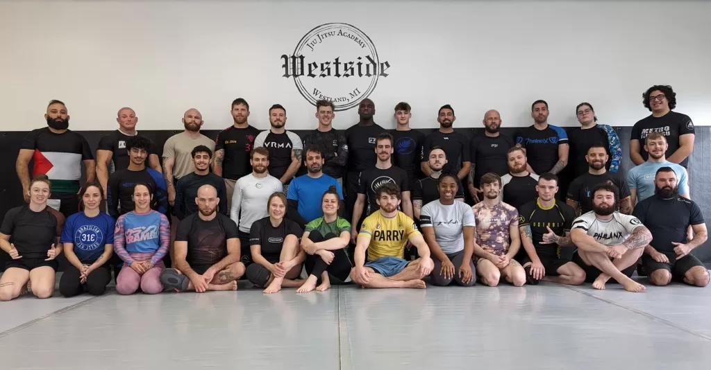 group of men and women of different ages sitting in rows on grey mats with black belts and academy logo in background