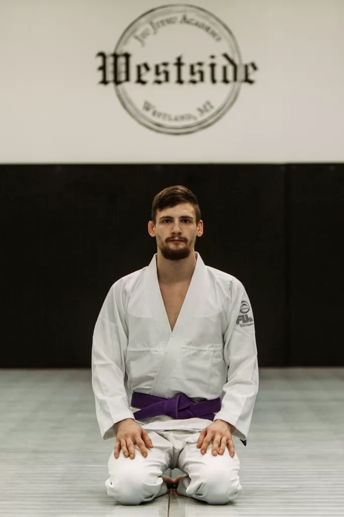 man kneeling on mats wearing a white gi with a purple belt, westside jiu jitsu logo above the mats on the wall