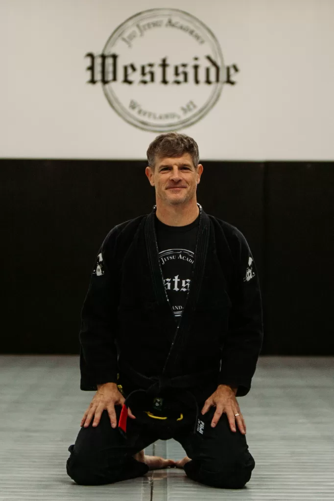 man kneeling on mats wearing a black gi and black belt with westside jiu jitsu academy logo on wall behind him