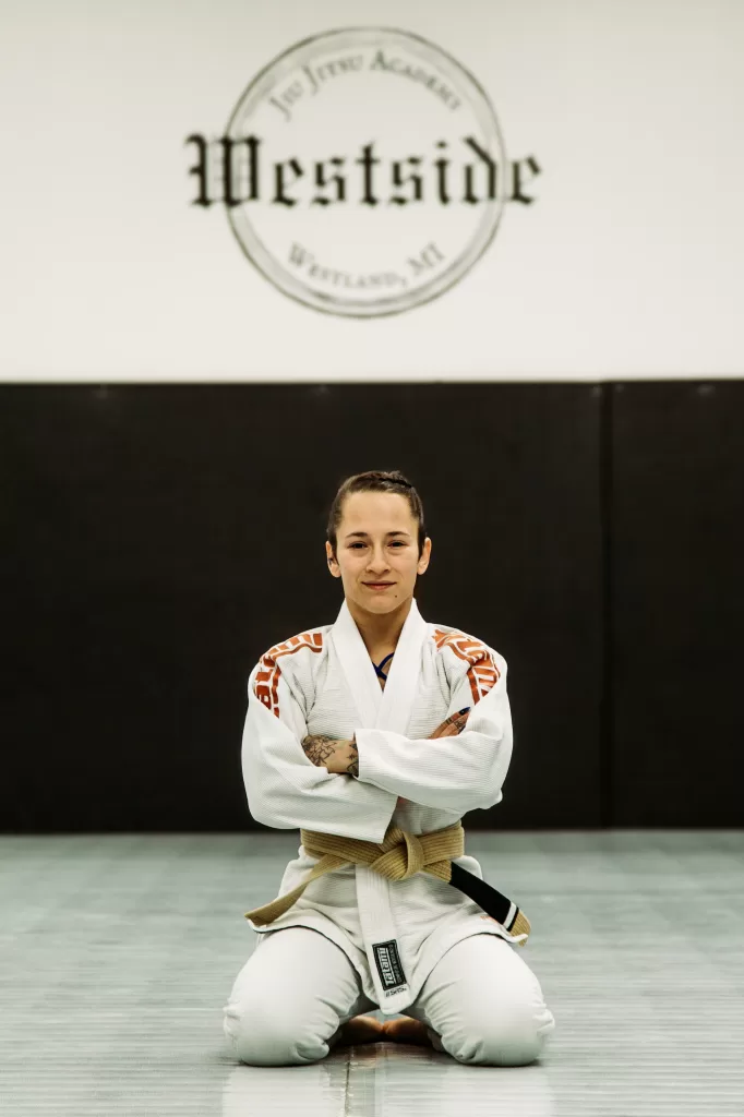 girl kneeling on mat arms crossed wearing a white gi and brown belt, westisde jiu jitsu logo on the wall in background