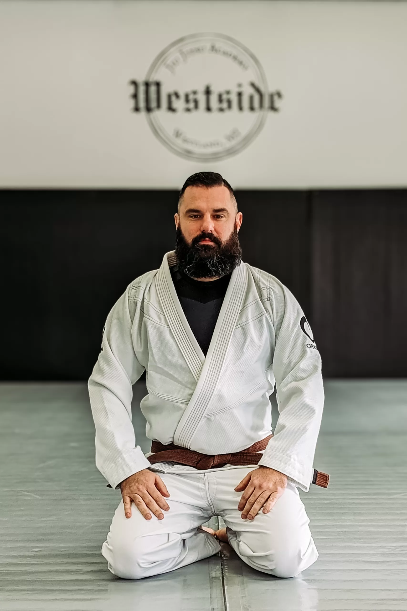 man in white gi and brown belt kneeling on grey mats with black mats and westside jiu jitsu academy logo in background