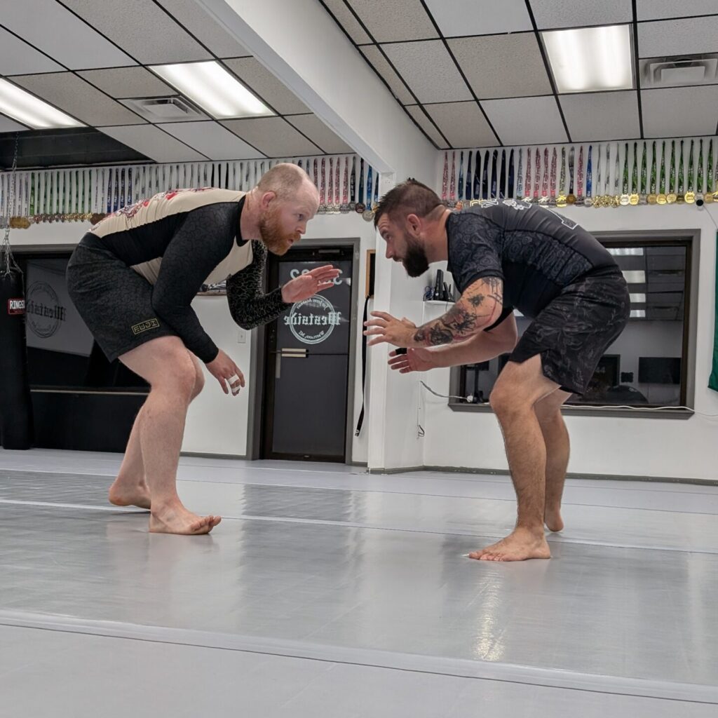 two men facing off in a grappling stance in center of mats, the background wall has two windows, a door, and medals hanging from the ceiling