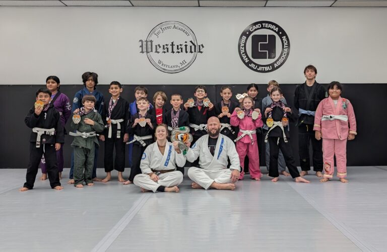 group photo of kids jiu jitsu students wearing gi's holding up medals with excited faces, coaches holding up giant silver medal sitting in front