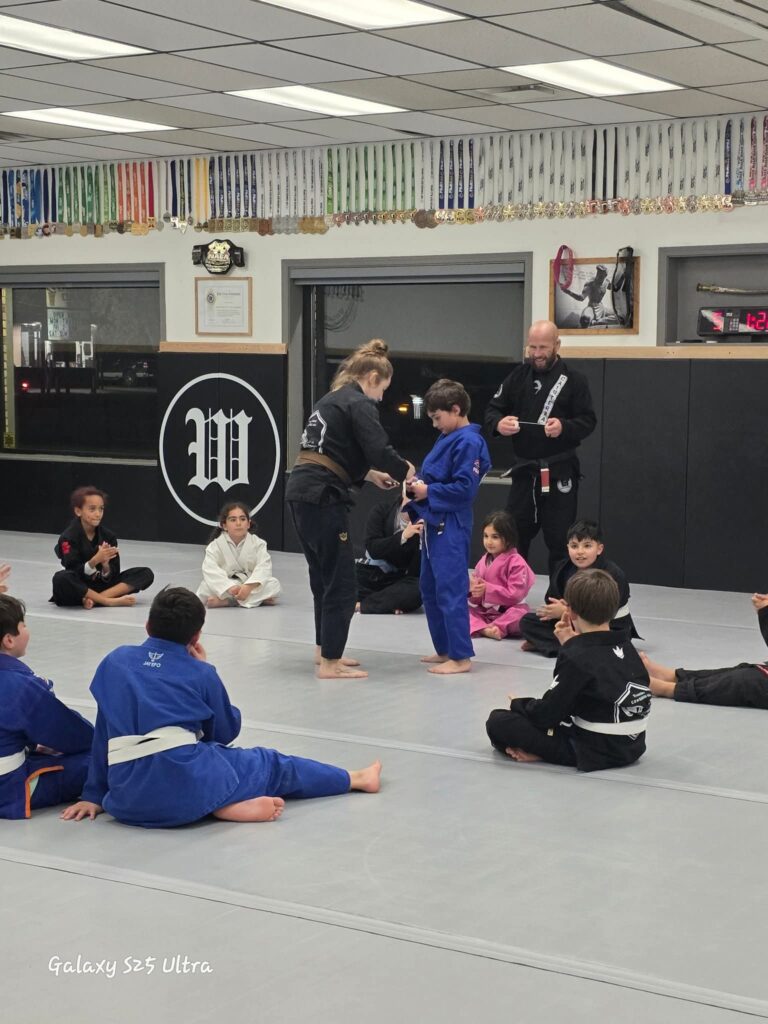 kids sitting in a circle wearing jiu jitsu gi's watching one of their peers be awarded a stripe by the instructors