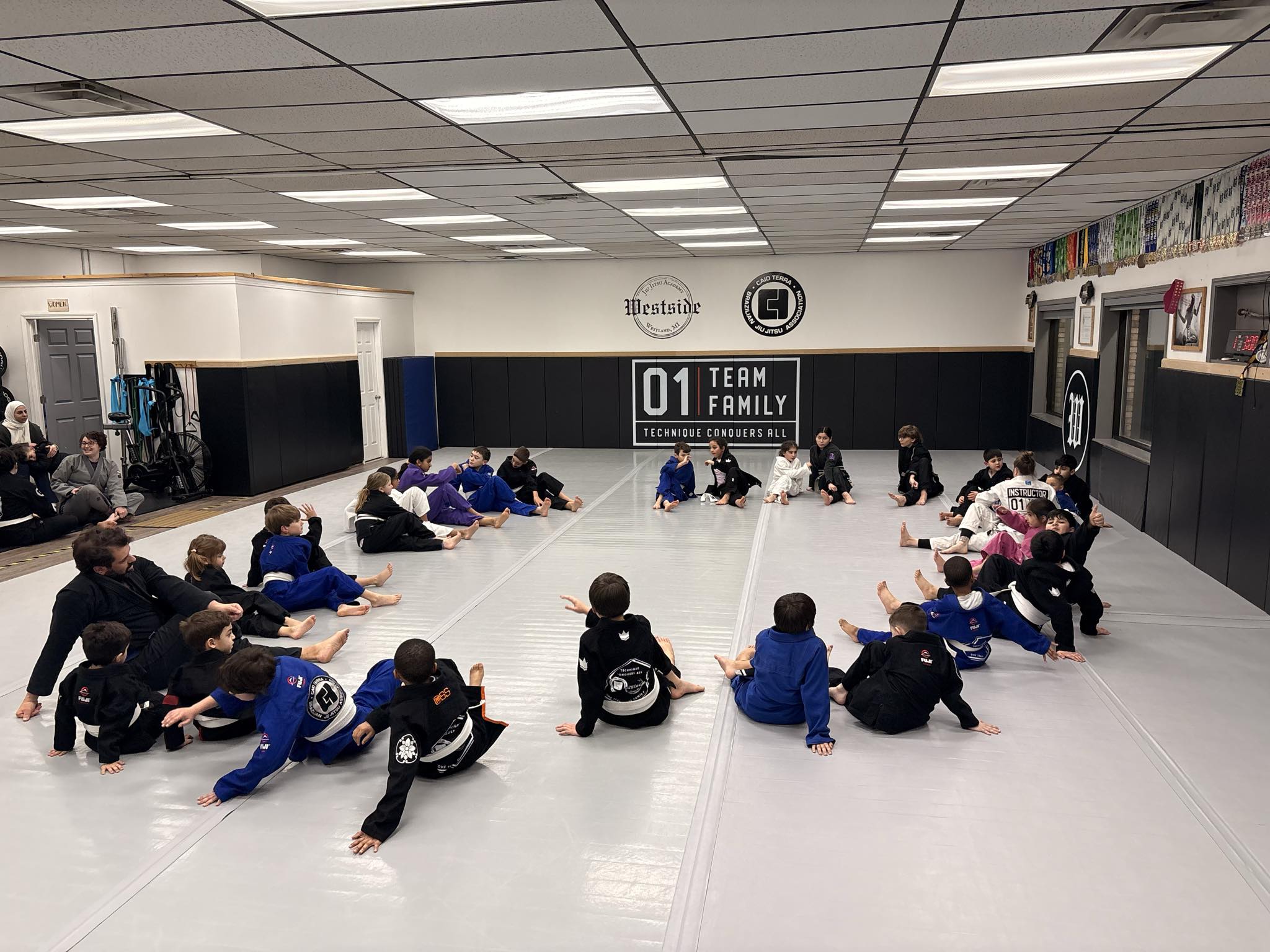 kids wearing jiu jitsu gi's sitting in a big circle on the mat stretching after class