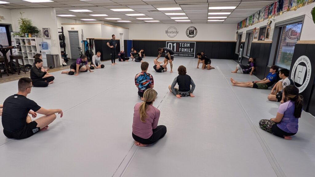 students sitting throughout the mat watching the instructor demonstrate a nogi jiu jitsu technique