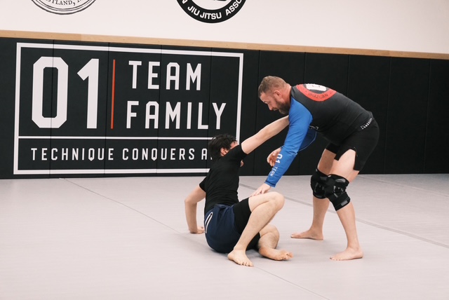 two students training jiu jitsu, one seated and one standing over him both reaching out to make contact